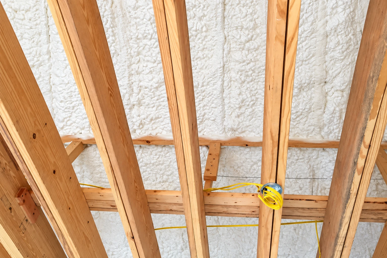 Exposed ceiling framing with white spray foam filling the cavities and yellow wiring running between joists.