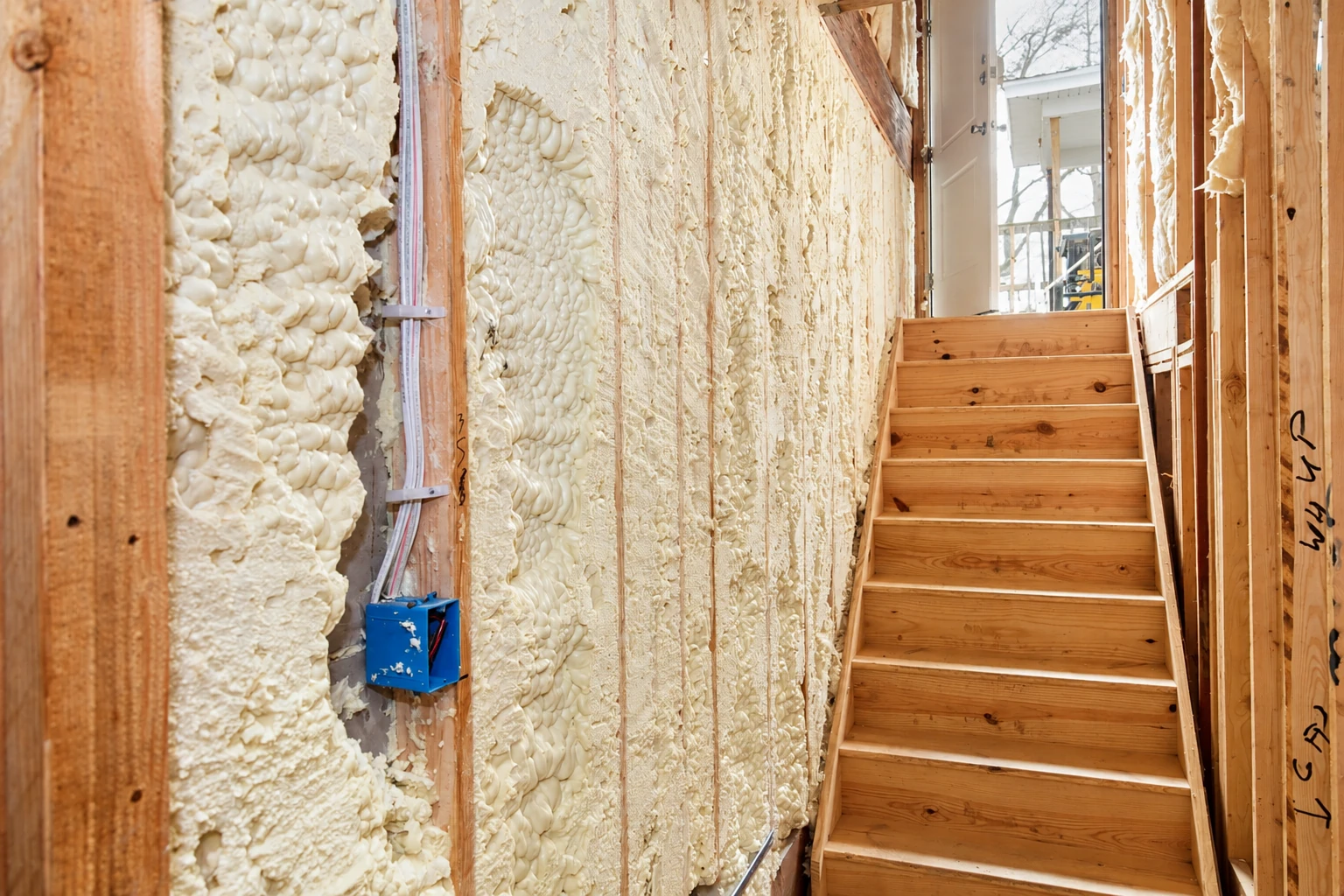 Wooden stairs beside exposed wall studs filled with uneven white spray foam insulation.