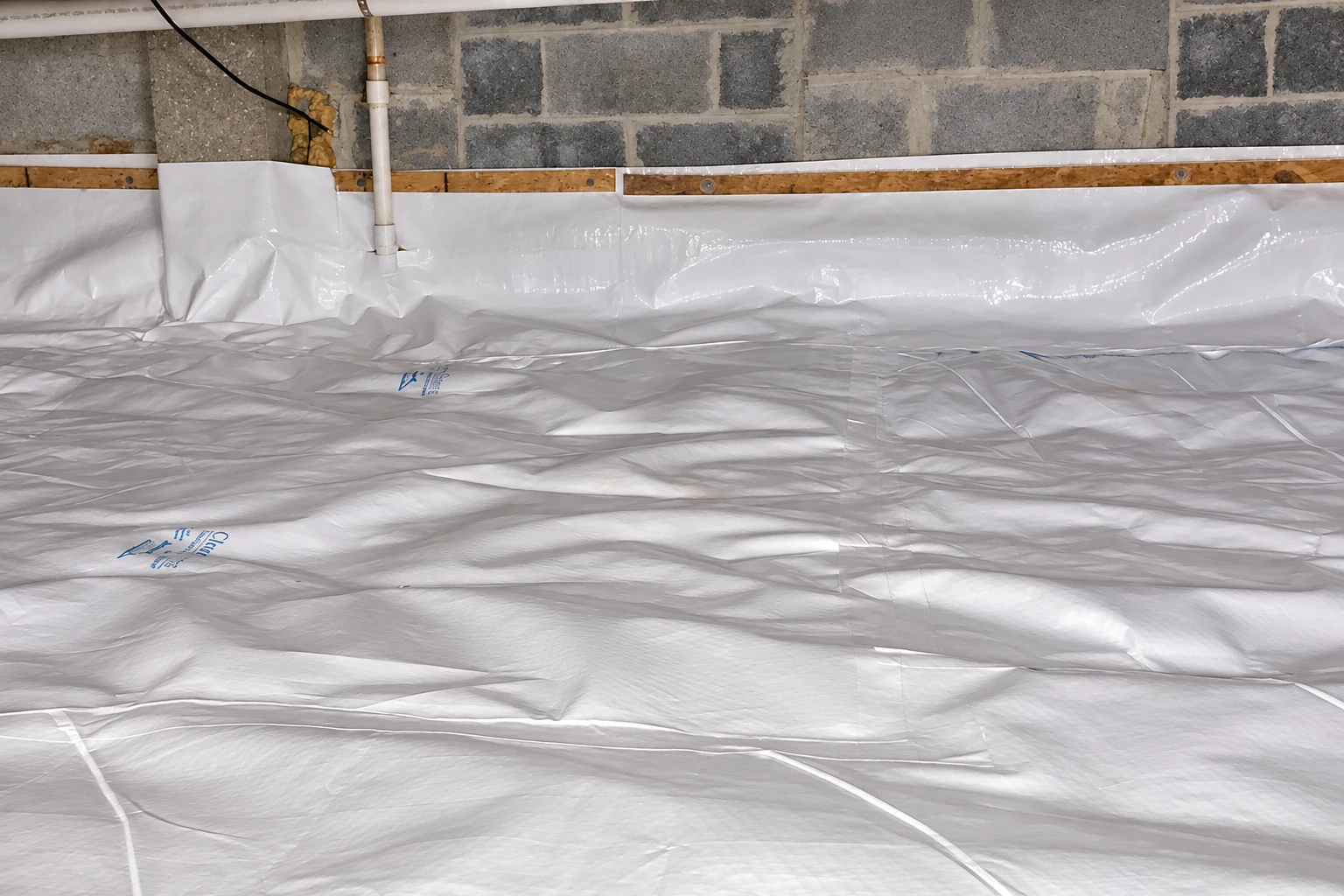 A crawl space floor is covered with white plastic vapor barrier beneath block walls and plumbing pipes.