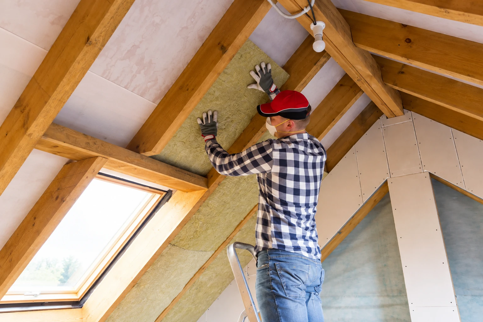 A worker presses yellow insulation batts between exposed rafters in a partly finished attic.