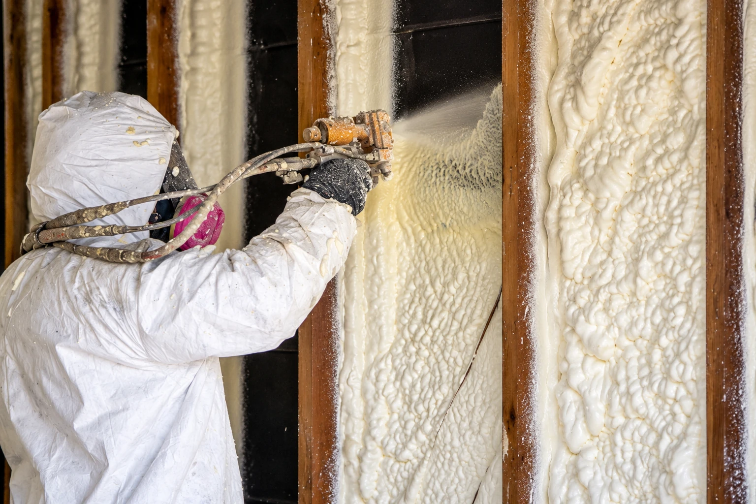 A worker in protective coveralls sprays expanding foam insulation between exposed wooden wall studs.